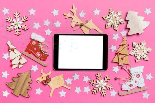 Top view of digital tablet, pink background decorated with festive toys and Christmas symbols reindeers and New Year trees. Holiday concept photo