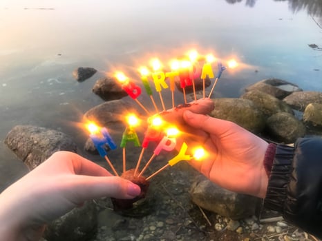 Burning happy birthday inscription made of holiday candles in the hands of a man and a woman opposite the water of the ocean lake river. Concept birthday celebration in nature, outdoors photo