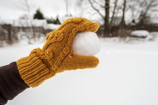 A hand in a yellow mitten holds a snowball, in the yard against the background of a snow-covered vegetable garden. Winter active games and fun. photo