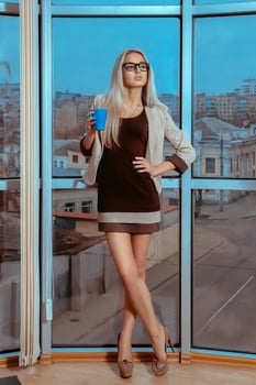 Vertical portrait of sexy blonde woman with cup of tea by the large window with city views photo