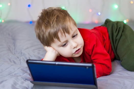 A cute boy is sitting on the bed and looking at a tablet. Funny smiling child. photo
