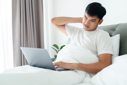 Asian man in white t-shirt laying on bed using laptop computer having neck pain. photo