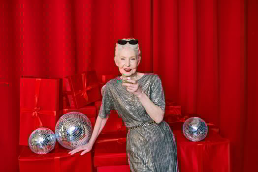 Senior stylish elegant woman with glass of sparkling wine on red background. Party, fashion, celebration, anti age concept photo