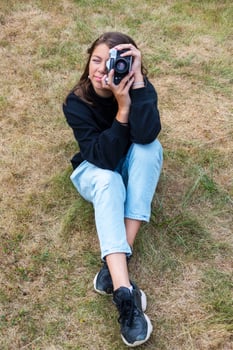 Cute teen girl with a camera, a girl taking pictures on a retro vintage camera on the grass in the park, a hobby concept photo