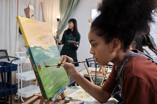African American girl concentrates on acrylic color picture painting on canvas with students group in art classroom, creative learning with talents and skills in elementary school studio education. photo