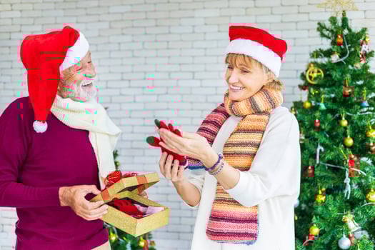 Happy smiling senior caucasian couple celebrating Christmas together while exchanging their present in happiness and excitement at home with red Santa hat and christmas tree photo