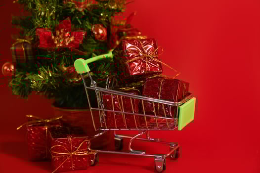 a Shopping cart full of gifts against red background near small christmas tree. Christmas or new year shopping photo