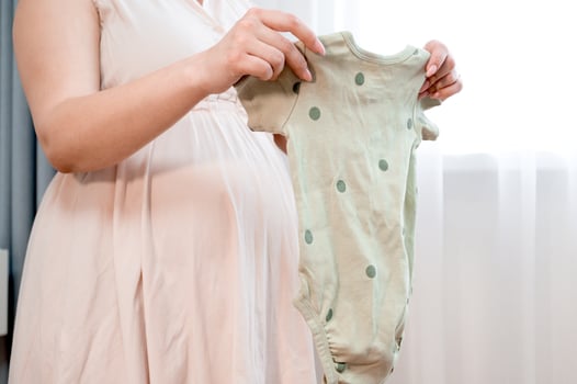 Pregnant woman standing against the window holding baby clothes. Preparing for parenthood. photo