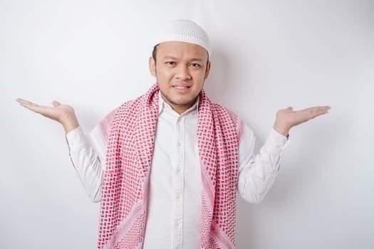A thoughtful young Muslim man shrugging his shoulders, gesturing confusion isolated by white background photo