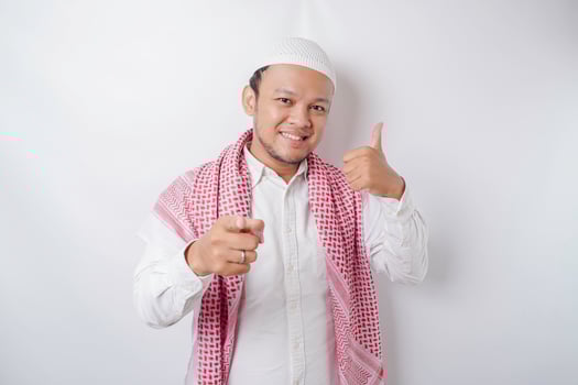 Excited Asian Muslim man gives thumbs up hand gesture of approval, isolated by white background photo