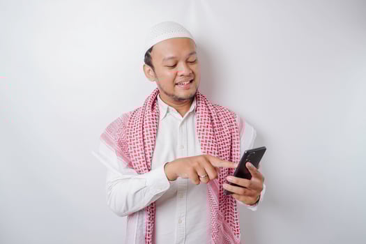 A portrait of a happy Asian Muslim man smiling while holding his phone, isolated by white background photo