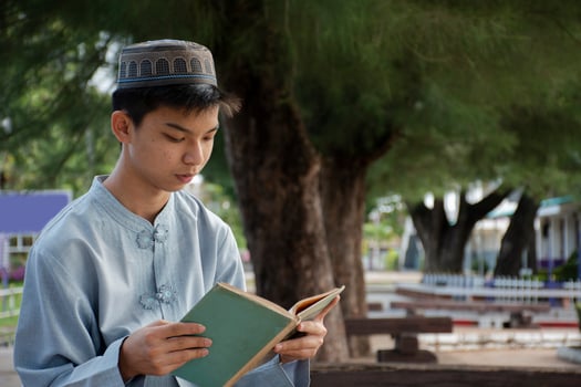 Young asian muslim boy wears hat, sitting in school park and reading his book in his free times before going back home, soft and selective focus. photo