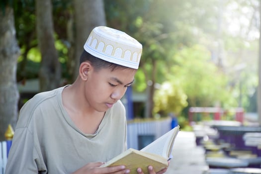 Young asian muslim boy wears hat, sitting in school park and reading his book in his free times before going back home, soft and selective focus. photo