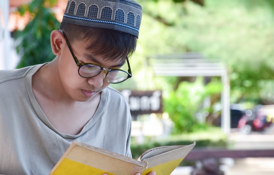 Young asian muslim boy wears eyeglasses, sitting in school park and reading his book in his free times before going back home, soft and selective focus. photo