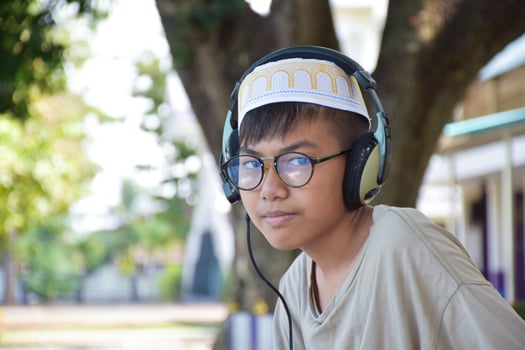 Portrait Young asian muslim boy in brown shirt and wears hat, holds headphone and sitting under the tree in the park, soft and selective focus. photo