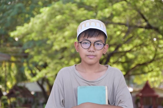 Young asian muslim boy wears eyeglasses, sitting in school park and reading his book in his free times before going back home, soft and selective focus. photo