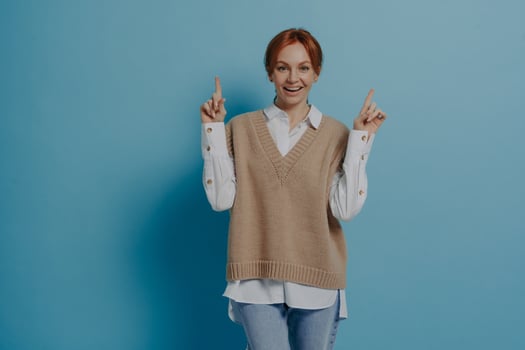 Happy red-haired woman showing up with forefingers at copy space and smiling, isolated on blue photo