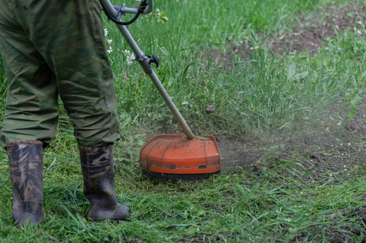 A close-up of a worker in protective clothing, gloves, rubber boots with a gas mower on the front lawn. A man mows grass with dandelions on a warm sunny spring day. photo