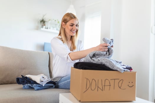 Happy young woman sit on couch stuck clothes in donation box at home, caring biracial female volunteer put apparel in carton package, donate to needy people, reuse, recycle concept photo