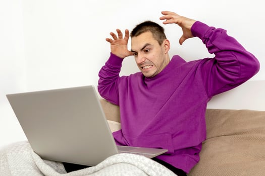 Angry and dissatisfied man with violet hoodie sitting on bed with laptop computer. Man using notebook to surf in internet, read news, watch movie, study or work online. Negative emotions, stress. photo