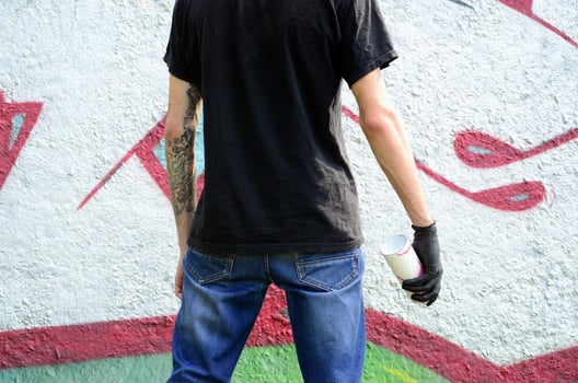 A young hooligan with a spray can stands against a concrete wall with graffiti paintings. Illegal vandalism concept. Street art photo