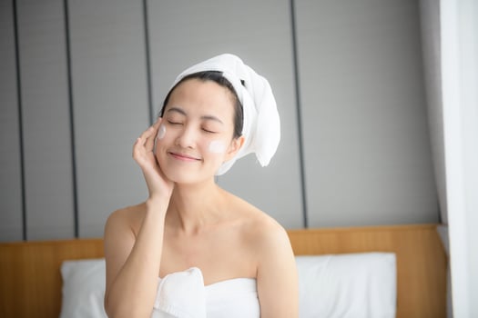 Happy young Asian woman applying face lotions while wearing a towel and touching her face. Daily makeup and skincare photo