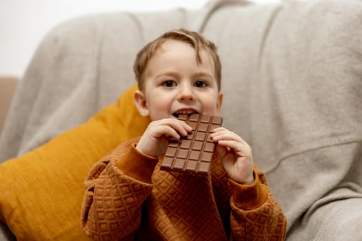 Little adorable boy sitting on the couch at home and eating chocolate bar. Child and sweets, sugar confectionery. Kid enjoy a delicious dessert. Preschool child with casual clothing. Positive emotion. photo
