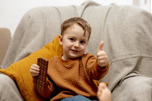 Little adorable boy sitting on the couch at home and eating chocolate bar. Child and sweets, sugar confectionery. Kid enjoy a delicious dessert. Preschool child with casual clothing. Positive emotion. photo