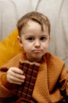 Little adorable boy sitting on the couch at home and eating chocolate bar. Child and sweets, sugar confectionery. Kid enjoy a delicious dessert. Preschool child with casual clothing. photo