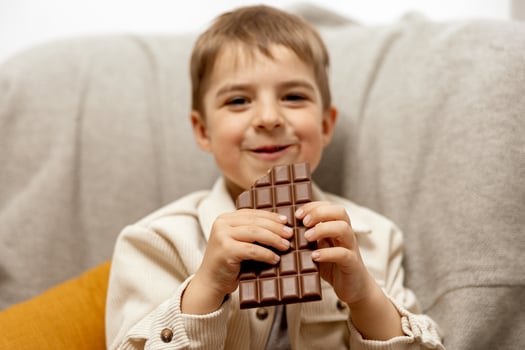 Little adorable boy sitting on the couch at home and eating chocolate bar. Child and sweets, sugar confectionery. Kid enjoy a delicious dessert. Preschool child with casual clothing. Positive emotion. photo
