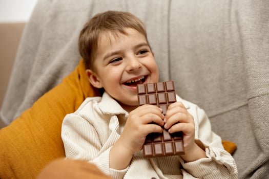 Little adorable boy sitting on the couch at home and eating chocolate bar. Child and sweets, sugar confectionery. Kid enjoy a delicious dessert. Preschool child with casual clothing. Positive emotion. photo