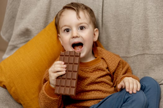Little adorable boy sitting on the couch at home and eating chocolate bar. Child and sweets, sugar confectionery. Kid enjoy a delicious dessert. Preschool child with casual clothing. Positive emotion. photo