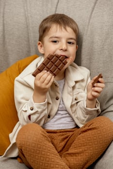 Little adorable boy sitting on the couch at home and eating chocolate bar. Child and sweets, sugar confectionery. Kid enjoy a delicious dessert. Preschool child with casual clothing. Positive emotion. photo