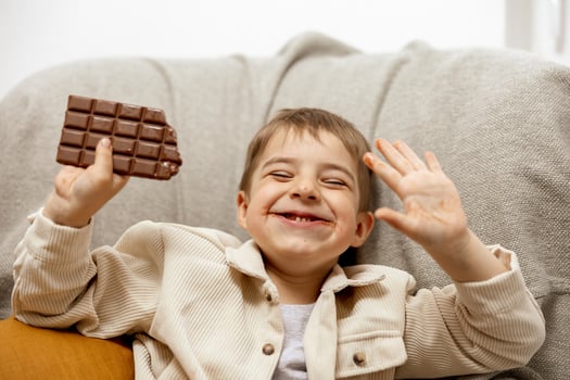 Little adorable boy sitting on the couch at home and eating chocolate bar. Child and sweets, sugar confectionery. Kid enjoy a delicious dessert. Preschool child with casual clothing. Positive emotion. photo