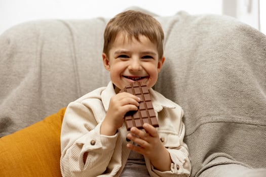 Little adorable boy sitting on the couch at home and eating chocolate bar. Child and sweets, sugar confectionery. Kid enjoy a delicious dessert. Preschool child with casual clothing. Positive emotion. photo