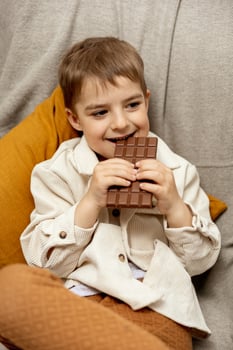 Little adorable boy sitting on the couch at home and eating chocolate bar. Child and sweets, sugar confectionery. Kid enjoy a delicious dessert. Preschool child with casual clothing. Positive emotion. photo