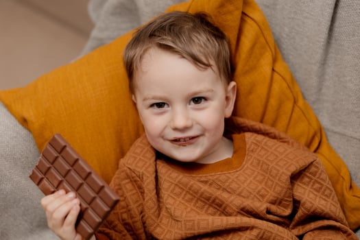 Little adorable boy sitting on the couch at home and eating chocolate bar. Child and sweets, sugar confectionery. Kid enjoy a delicious dessert. Preschool child with casual clothing. Positive emotion. photo
