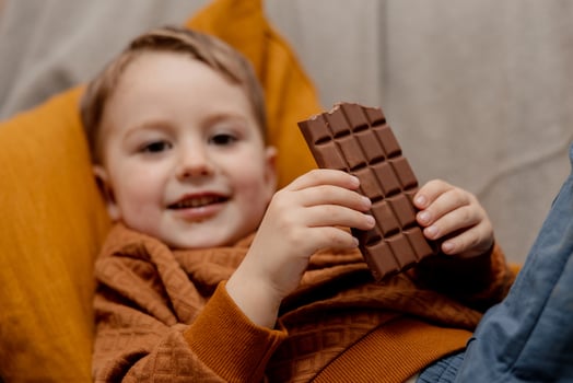 Little adorable boy sitting on the couch at home and eating chocolate bar. Child and sweets, sugar confectionery. Kid enjoy a delicious dessert. Preschool child with casual clothing. Positive emotion. photo