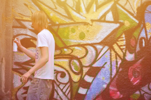 Photo of a young guy in denim shorts and a white shirt. The guy draws on the graffiti wall a drawing with aerosol paints of various colors. The concept of hooliganism and damage to property