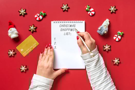 female hand holds pen and writing Christmas shopping list, gift ideas on white notepad on red background Top view Flat lay Holiday card Woman making Christmas presents list, Planning, buying concept photo