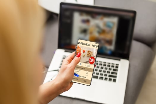 Woman using laptop and smartphone in cafe holding phone with app delivery food screen photo
