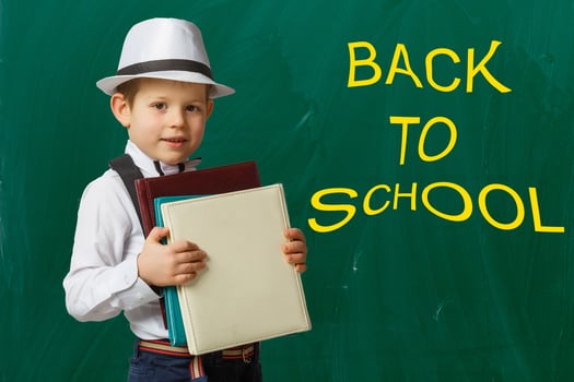 Back to school. Funny little boy in glasses pointing up on blackboard. Child from elementary school with book and bag. Education. photo