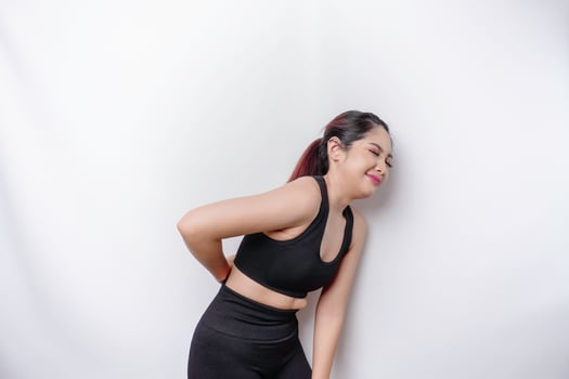 Tired upset sporty Asian woman wearing sportswear is suffering from back pain, muscle spasm at workplace. Fatigue, deadline, pain and incorrect posture photo