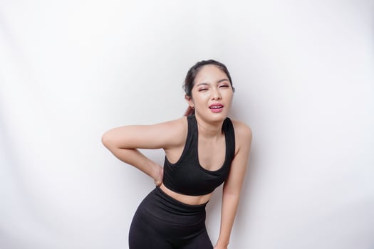 Tired upset sporty Asian woman wearing sportswear is suffering from back pain, muscle spasm at workplace. Fatigue, deadline, pain and incorrect posture photo