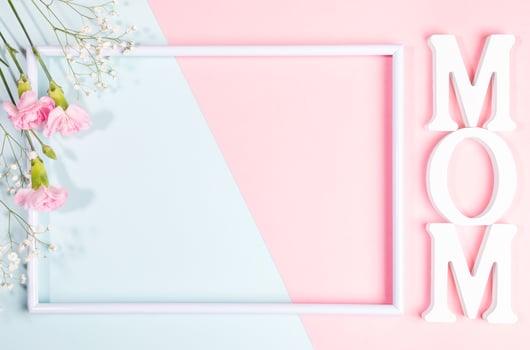 Empty white frame, pink carnations and gypsophila flowers, white letters MOM on pink- blue backdrop. photo