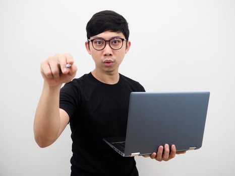 Asian man with glasses holding laptop point pen at you serious face on white background photo