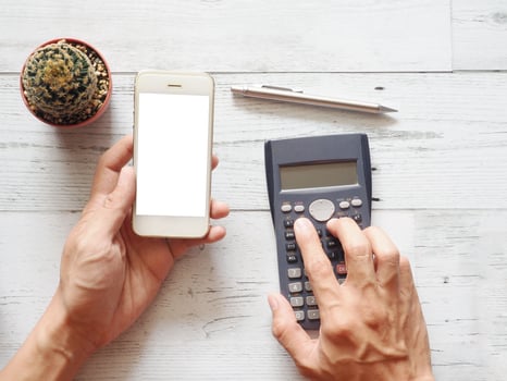 Hand holding cellphone white screen and using calculator with cactus and silver pen on white wood table nature shadow and sunlight top view and space business concept photo