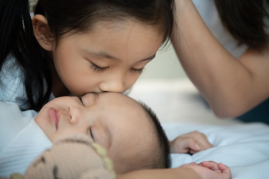 An older sister hugs her newborn little sister while she sleeps. In the white bedroom photo