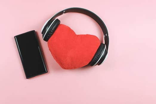 flat lay of red heart pillow coverd with headphones and blank black screen mobile phone on pink background. Love songs or podcast concept. photo