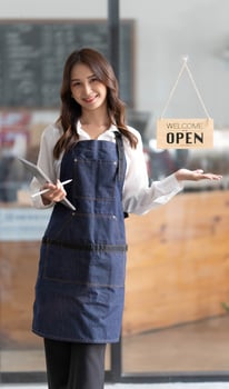 Beautiful asian young barista woman in apron holding tablet and standing in front of the door of cafe with open sign board. Business owner startup concept. photo